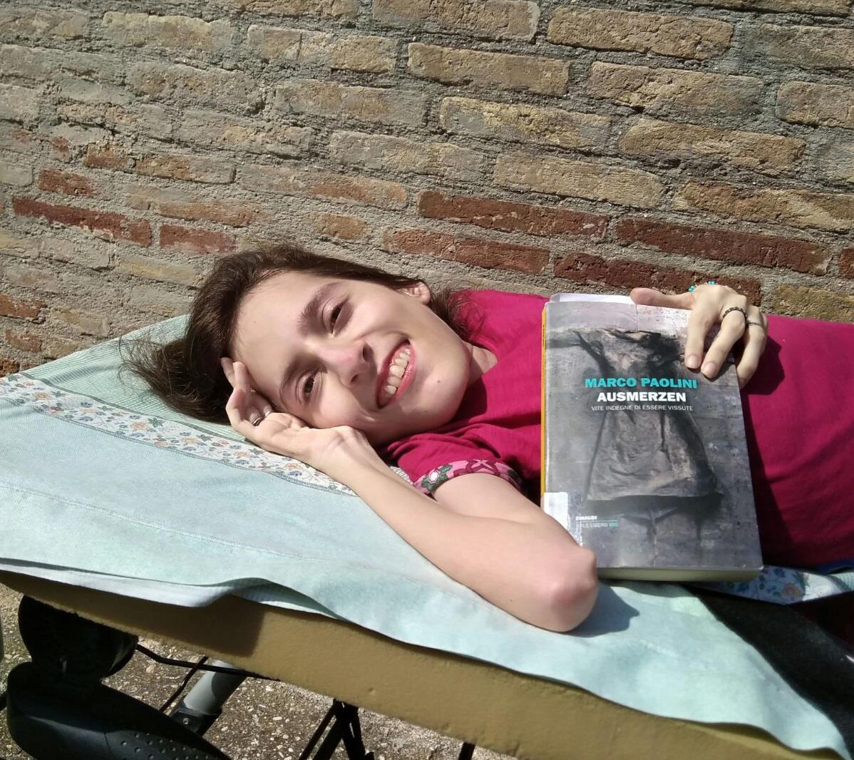 Elena è distesa, all'aperto, davanti a un muro di mattoni. Indossa una maglietta fucsia, mostra il libro di Marco Paolini, "Ausmerzen - Vite indegne di essere vissute".