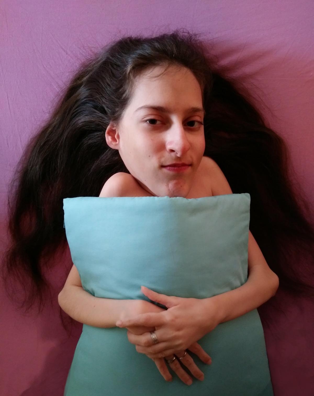Elena ha un cuscino azzurro tenuto davanti a sé con le braccia incrociate; ha i capelli lunghi castani e accenna un sorriso. Lo sfondo è rosa.
