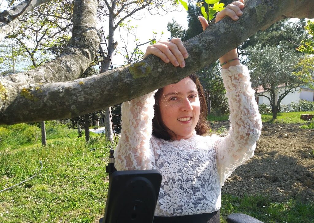 M. Chiara è in giardino, indossa una maglia bianca, appoggia le mani su un ramo e sorride.