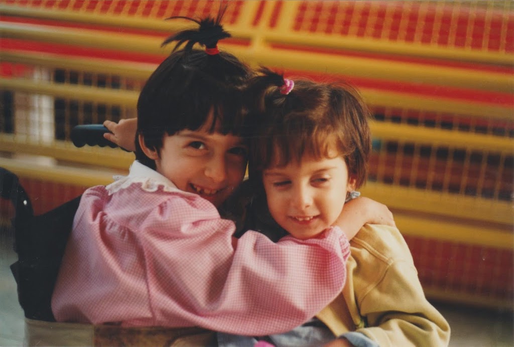 Chiara a Elena a circa 7 e 3 anni, a scuola. Elena è in braccio a Chiara che indossa un grembiule rosa. Entrambe hanno i capelli tirati su in un ciuccetto. Chiara sorride guardando l'obiettivo, Elena guarda da un'altra parte e sorride con gli occhi semichiusi.