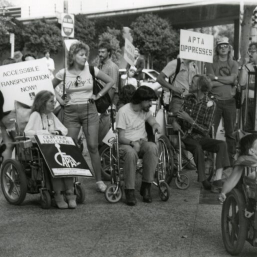 foto in bianco e nero di membri di ADAPT, tra cui Wade Blank, a una protesta per l'accessibilità degli autobus. Ci sono varie persone in carrozzina e alcune in piedi tra cui un poliziotto di spalle. Le persone disabili hanno dei cartelli di protesta.