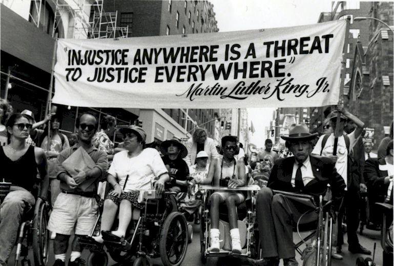 Foto d’epoca di un corteo di protesta di attivisti in carrozzina e non, bianchi e neri, con il cartello “Injustice anywhere is a threat to justice everywhere” (Martin Luther King Jr.)