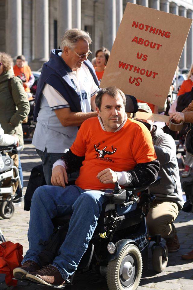 un uomo in carrozzina elettrica con indosso la maglietta arancione della Freedom Drive sfila con il cartello “Nothing about us without us”