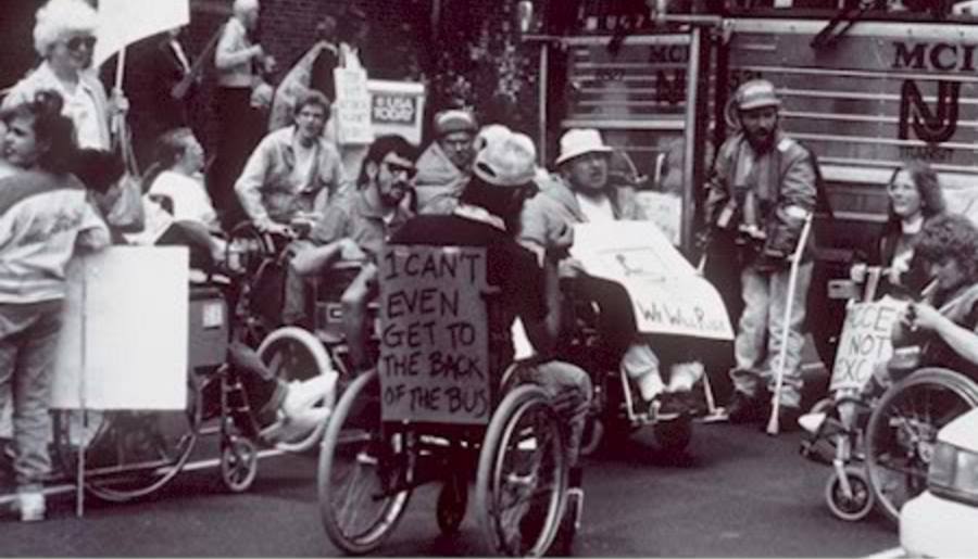 Una folla di persone disabili tiene dei cartelli di protesta. In primo piano, sullo schienale della carrozzina di un uomo, un cartello dice: "I can't even get to the back of the bus".
