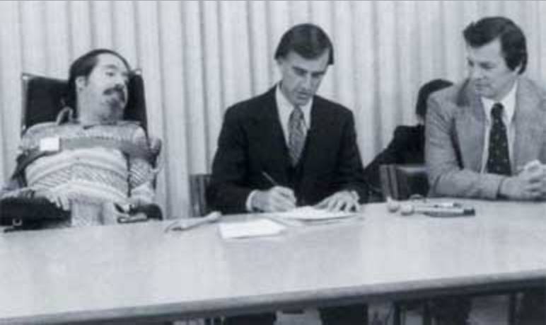 A un tavolo di lavoro sono seduti Ed Roberts e due uomini in giacca e cravatta, uno dei quali sta firmando un documento.