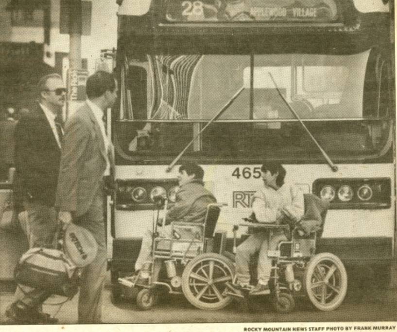 Due disabili in carrozzina bloccano un autobus stando sulla strada e impedendogli di partire. Un passante osserva la scena.