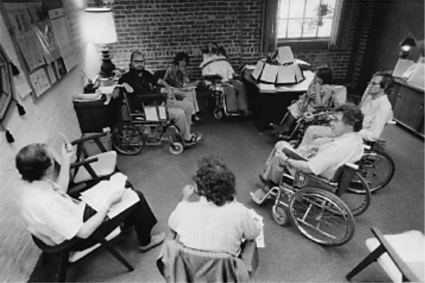 Attivisti disabili riuniti in cerchio in un locale, Ed Roberts sullo sfondo.