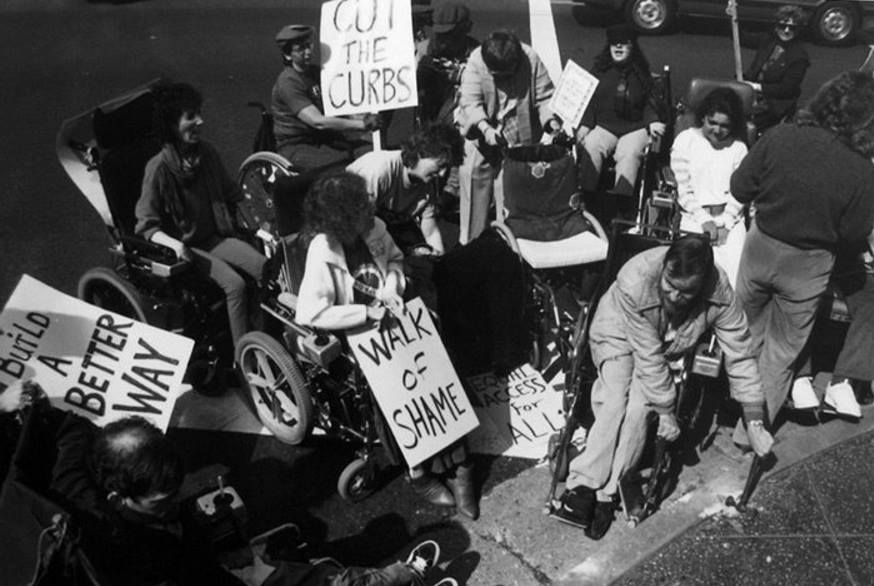 Un sit-in di protesta di disabili in carrozzina con i seguenti cartelli: “Build a better way”, “Cut the curbs”, “Walk of shame”, “Equal access for all”.