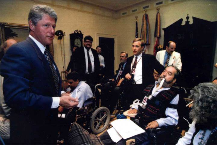 in primo piano c’è Bill Clinton, che indossa una giacca blu. Un gruppo di persone, tra cui Ed, lo osserva.