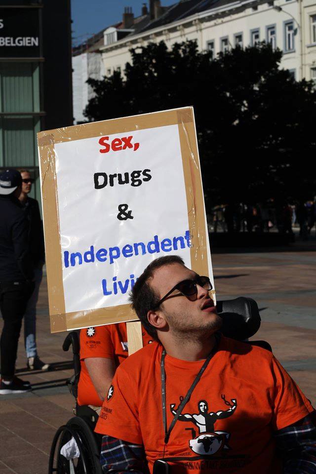 Un ragazzo in carrozzina con indosso la maglietta arancione della Freedom Drive e gli occhiali da sole sfila con il cartello “Sex, drugs and Independent Living”.