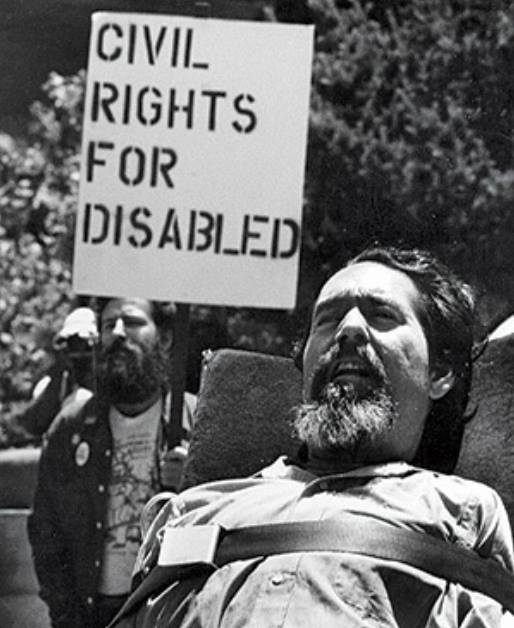 Ed Roberts durante una protesta. Un uomo dietro di lui regge un cartello con scritto "civil rights for disabled". Ed ha la barba brizzolata, indossa una camicia e ha un'espressione di orgoglio sul viso.