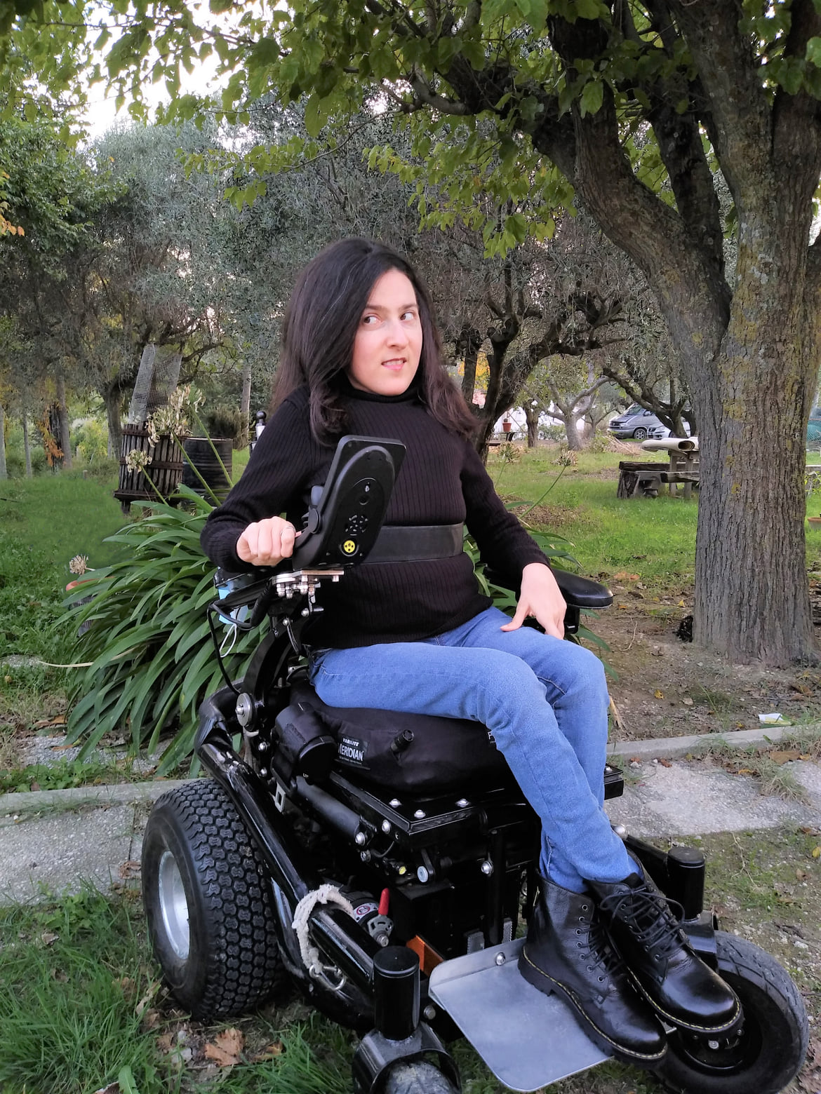 Maria Chiara indossa una maglia nera a collo alto, jeans blu e anfibi neri e guarda di lato. Sullo sfondo, un giardino.