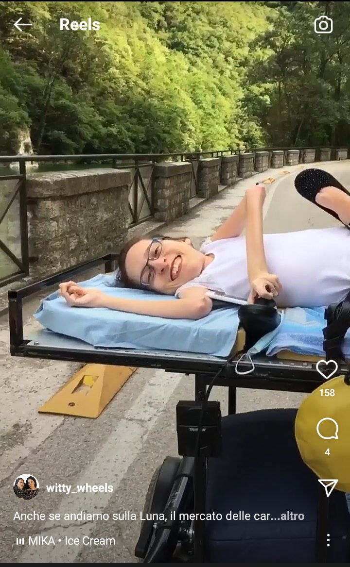 Screenshot di un reel da Instagram: Elena sorridente sulla sua carrozzina nera. Sullo sfondo, una strada e degli alberi